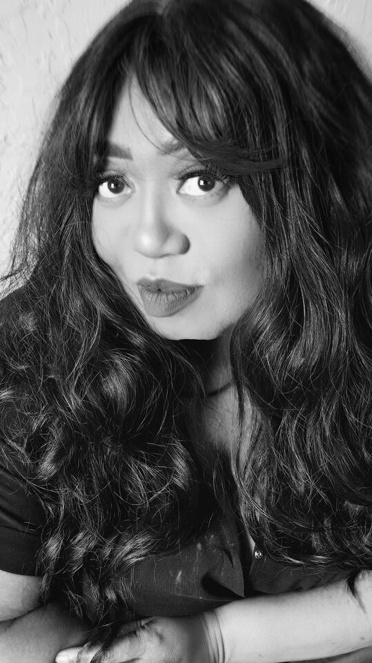 Nicole Gotfredson | Nicole in Practice | The Memorist | The Nicole Method Black and white close-up portrait of a woman with long, wavy hair and bangs, resting her arms forward with a soft, playful expression.